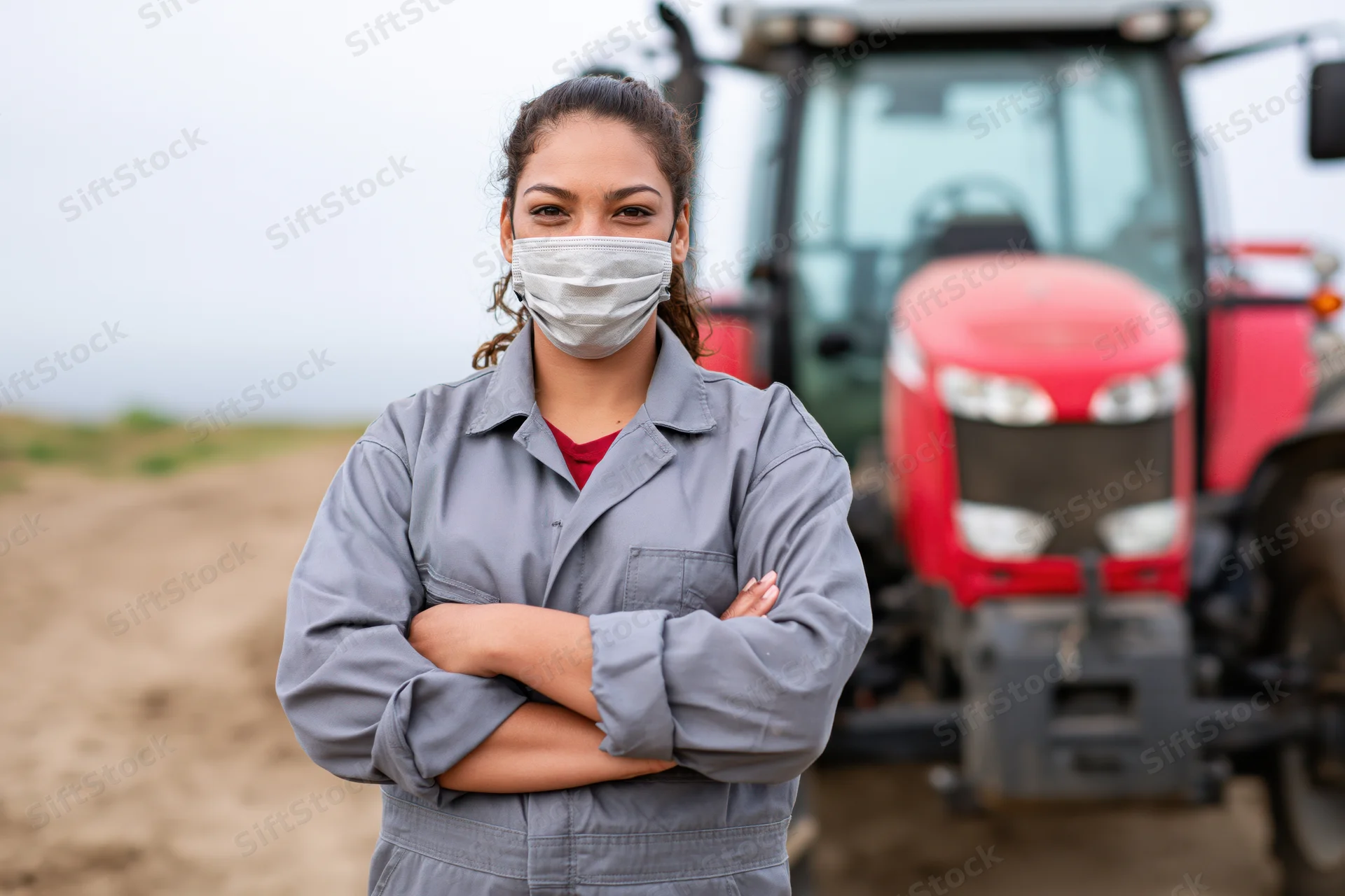 Woman in gray coveralls standing with arms crossed in front of red tractor on farm field