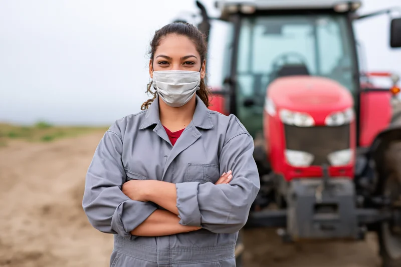 Woman in gray coveralls standing with arms crossed in front of red tractor on farm field