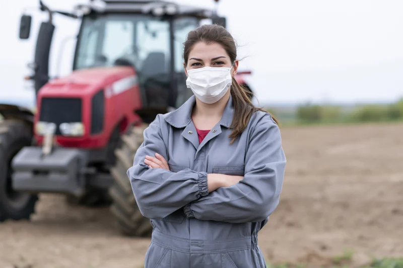 Woman in gray coveralls with arms crossed standing in a field beside a red tractor