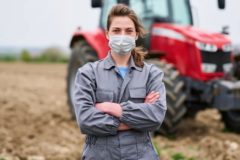 Woman in gray coveralls standing with arms crossed in front of red tractor on plowed field