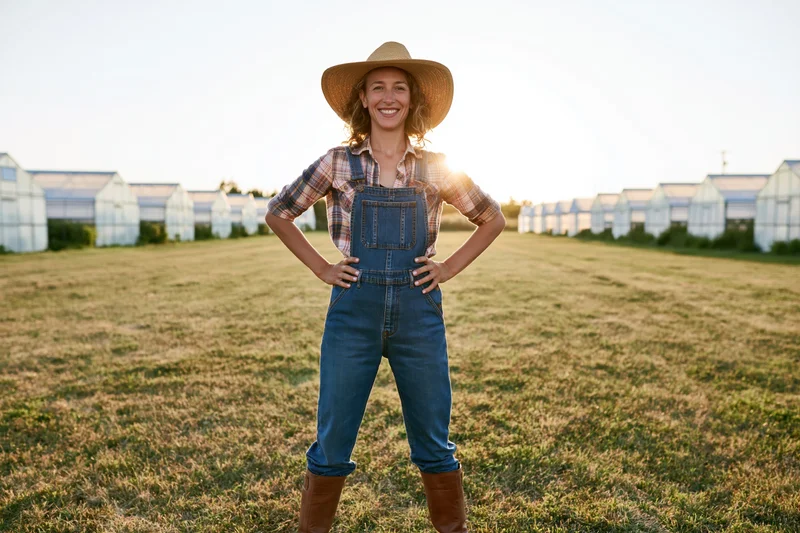 Woman in overalls and cowboy hat standing confidently with hands on hips in a field of greenhouses