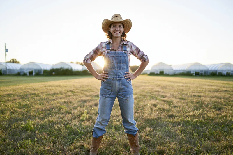 Woman in overalls and cowboy hat standing confidently with hands on hips in a grassy field at sunset near greenhouses