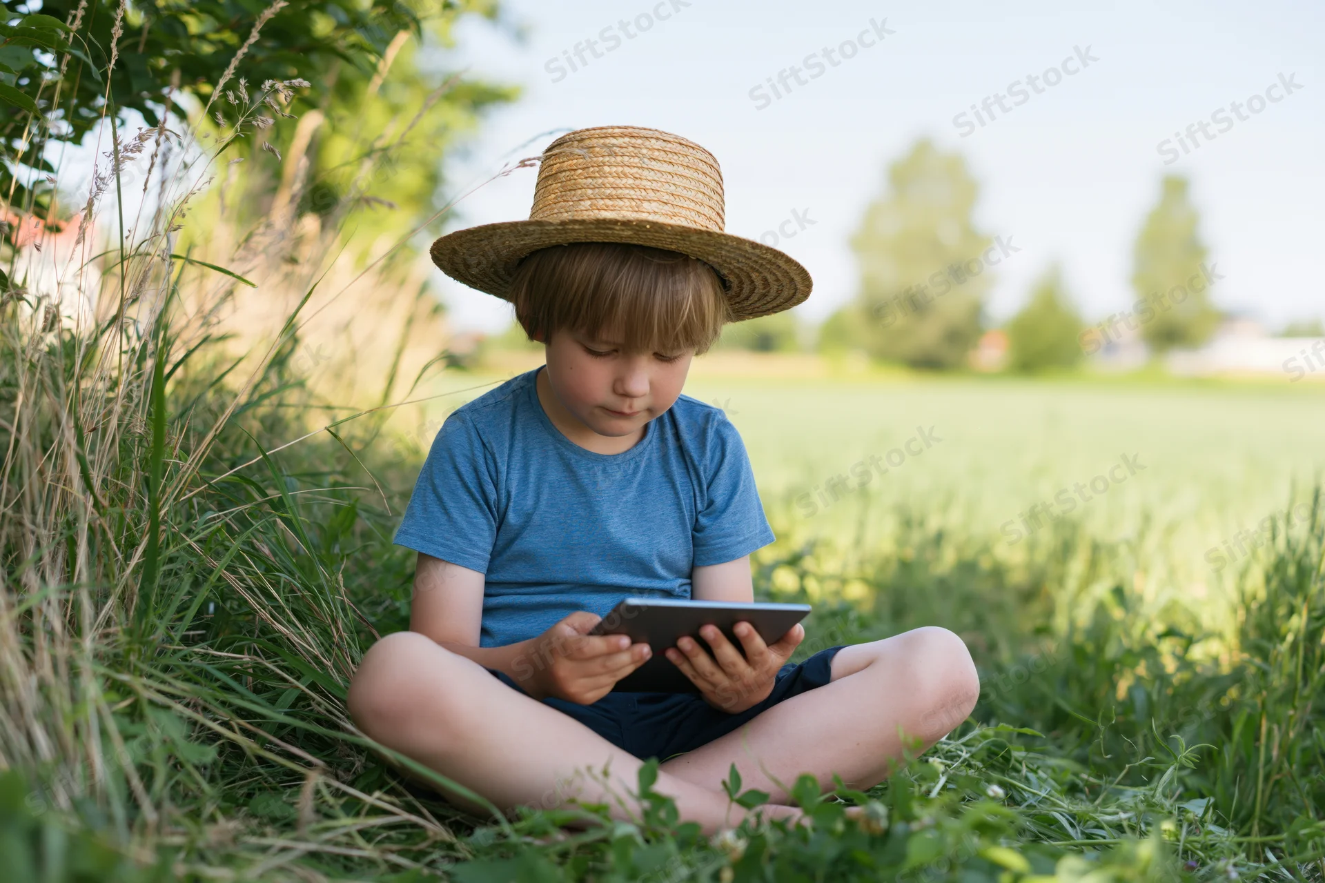 Young boy sitting crosslegged in grass using tablet outdoors