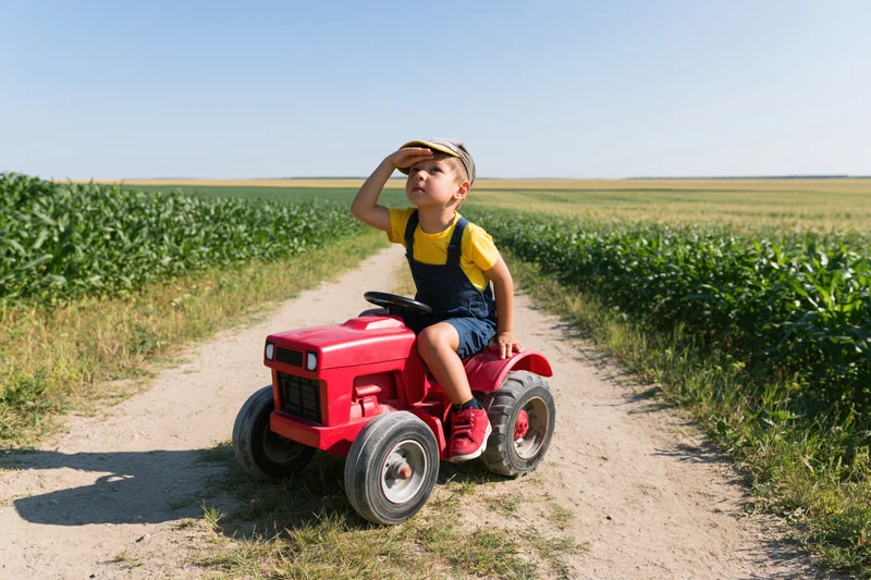 Young boy riding red toy tractor on dirt path through cornfield