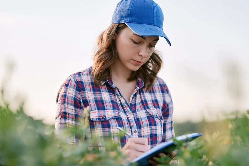 Young woman in plaid shirt and blue cap writing on clipboard in a green field