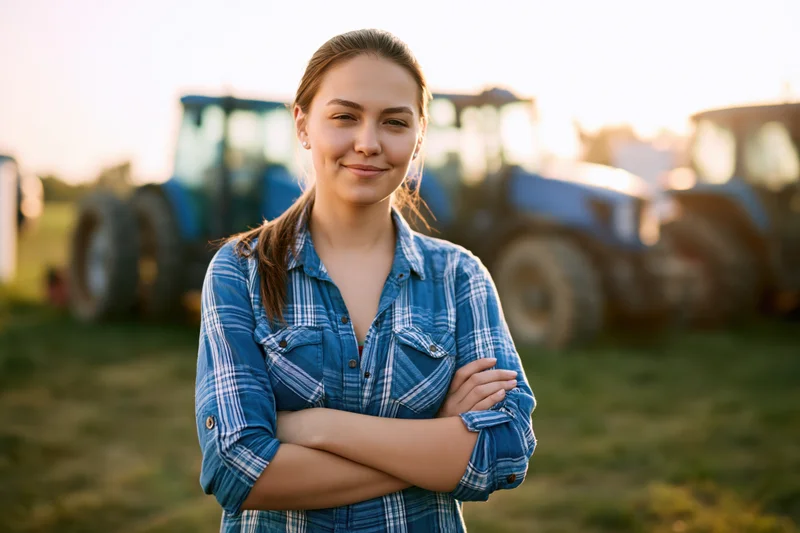 Young woman standing with arms crossed in a farm field at sunset, tractors in background