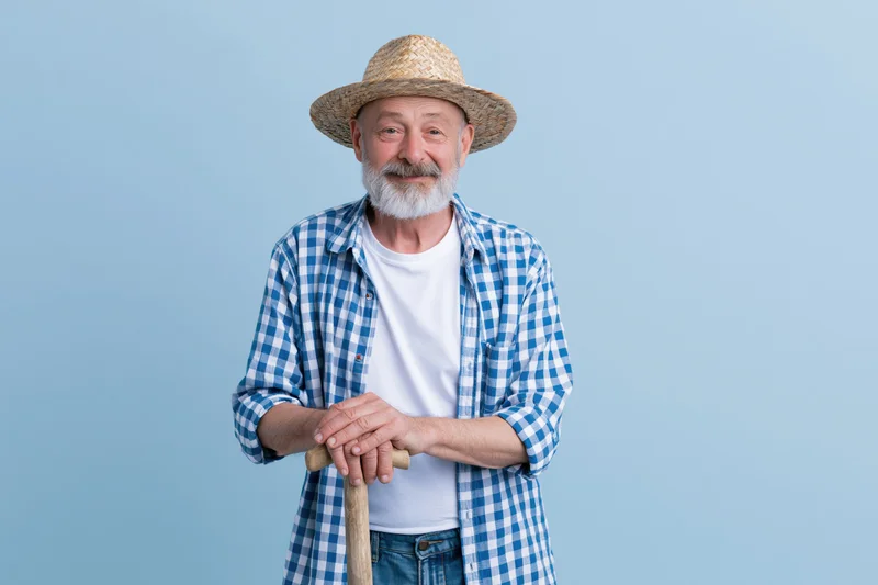 Senior man in straw hat and checkered shirt holding shovel, smiling in studio against light blue background