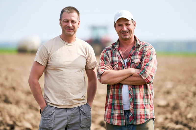 Two farmers standing together in a tilled field with a tractor in the background