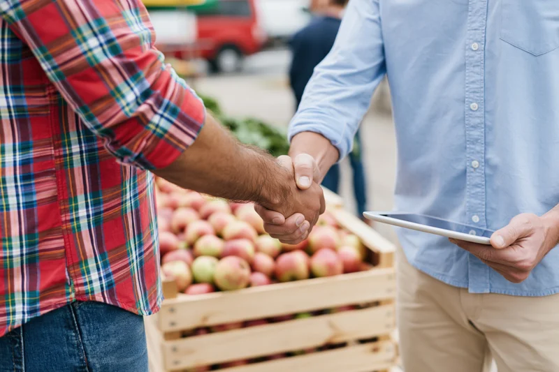 Two men shaking hands beside a wooden crate of apples at an outdoor market