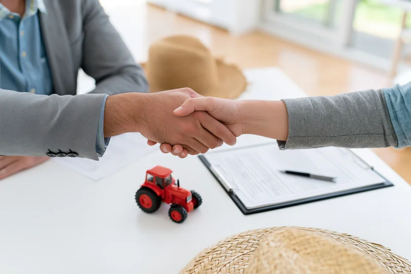 Two people shaking hands over a table with a red toy tractor and documents, indicating a farm-related agreement