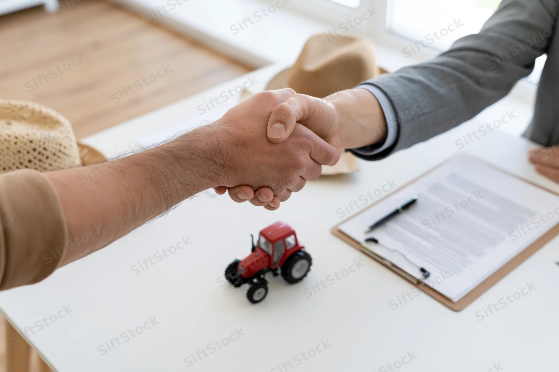 Two men shaking hands over a desk with a red toy tractor and contract clipboard in an office setting