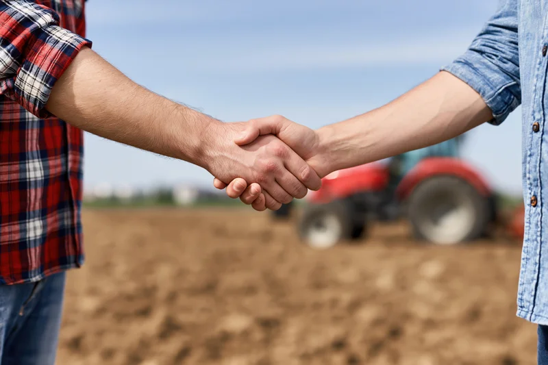 Two men shaking hands in a plowed field with a red tractor in the background