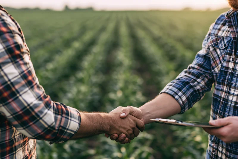 Two men in plaid shirts shaking hands in a crop field holding a tablet