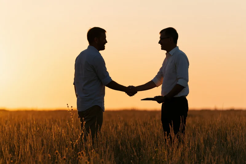 Two men shaking hands in a wheat field at sunset