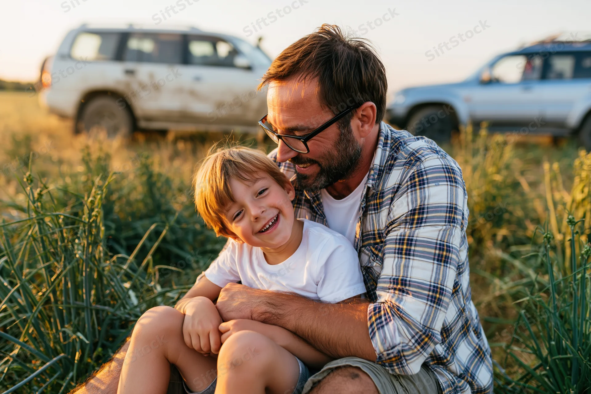 Father hugging his smiling son in a grassy field at sunset with SUVs in the background