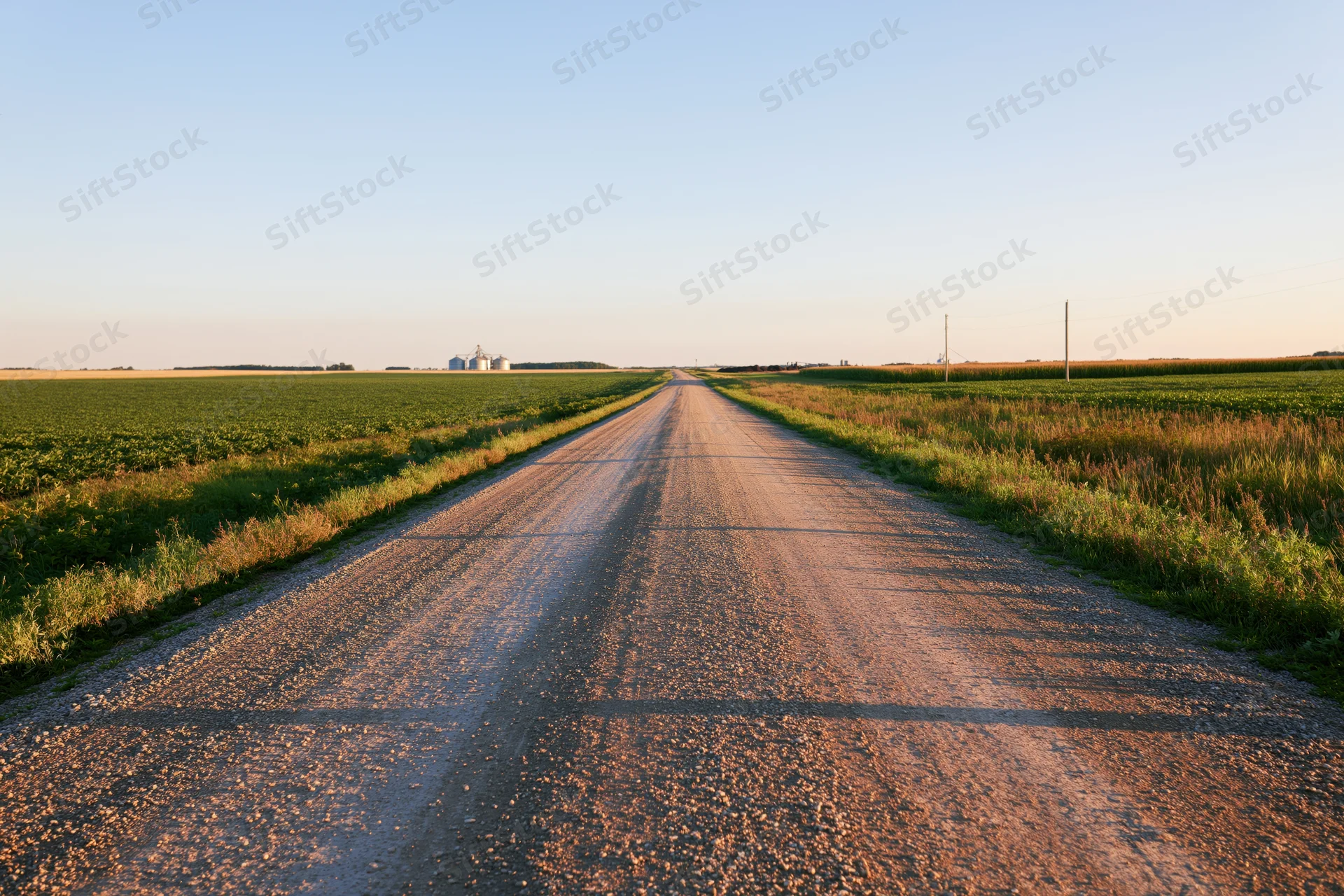 Straight gravel road stretching through flat farmland toward distant farm buildings at sunset