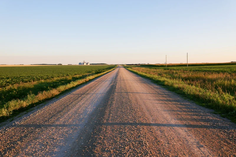 Straight gravel road stretching through flat farmland toward distant farm buildings at sunset
