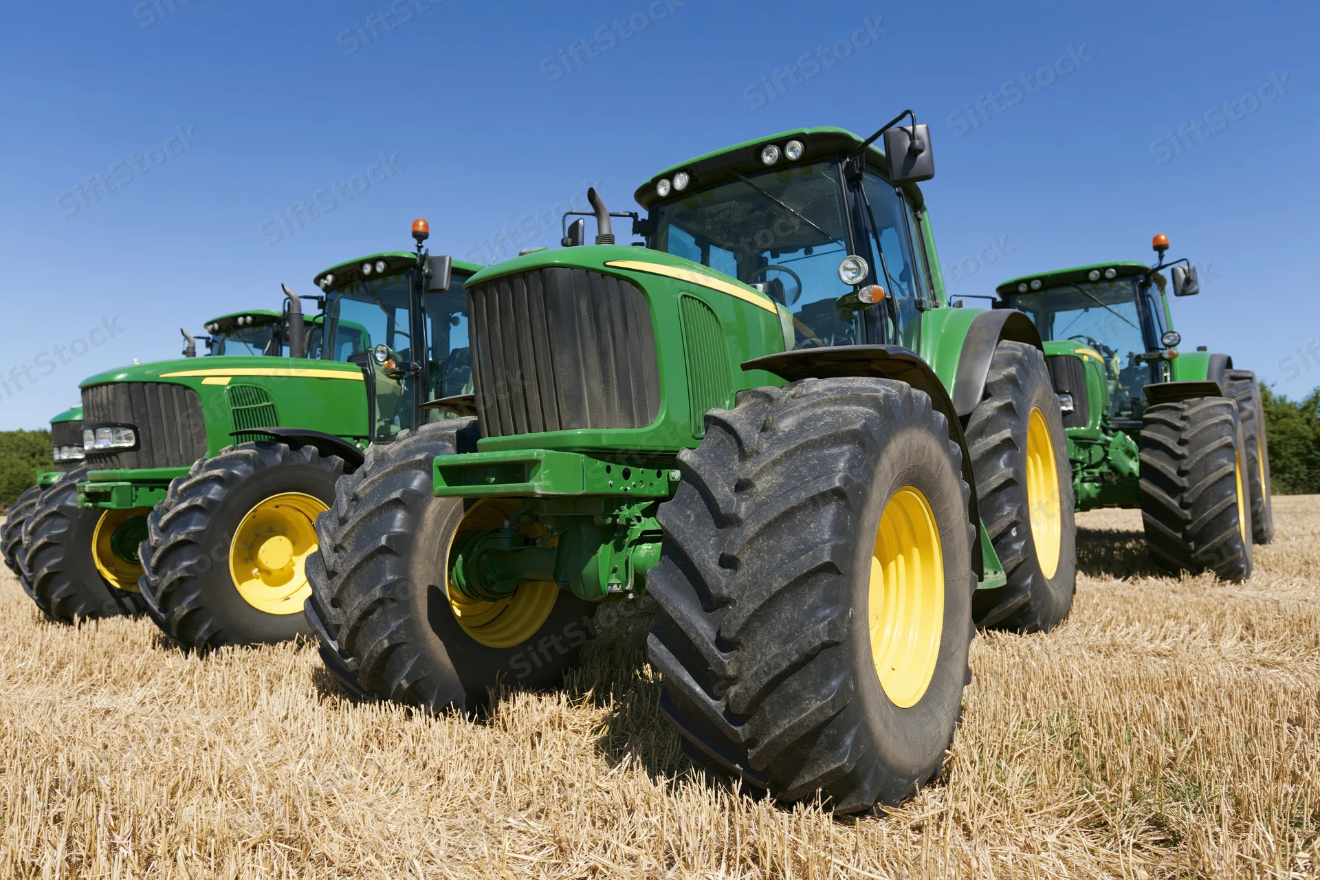 Three green and yellow tractors parked in a row on a harvested field under a clear blue sky