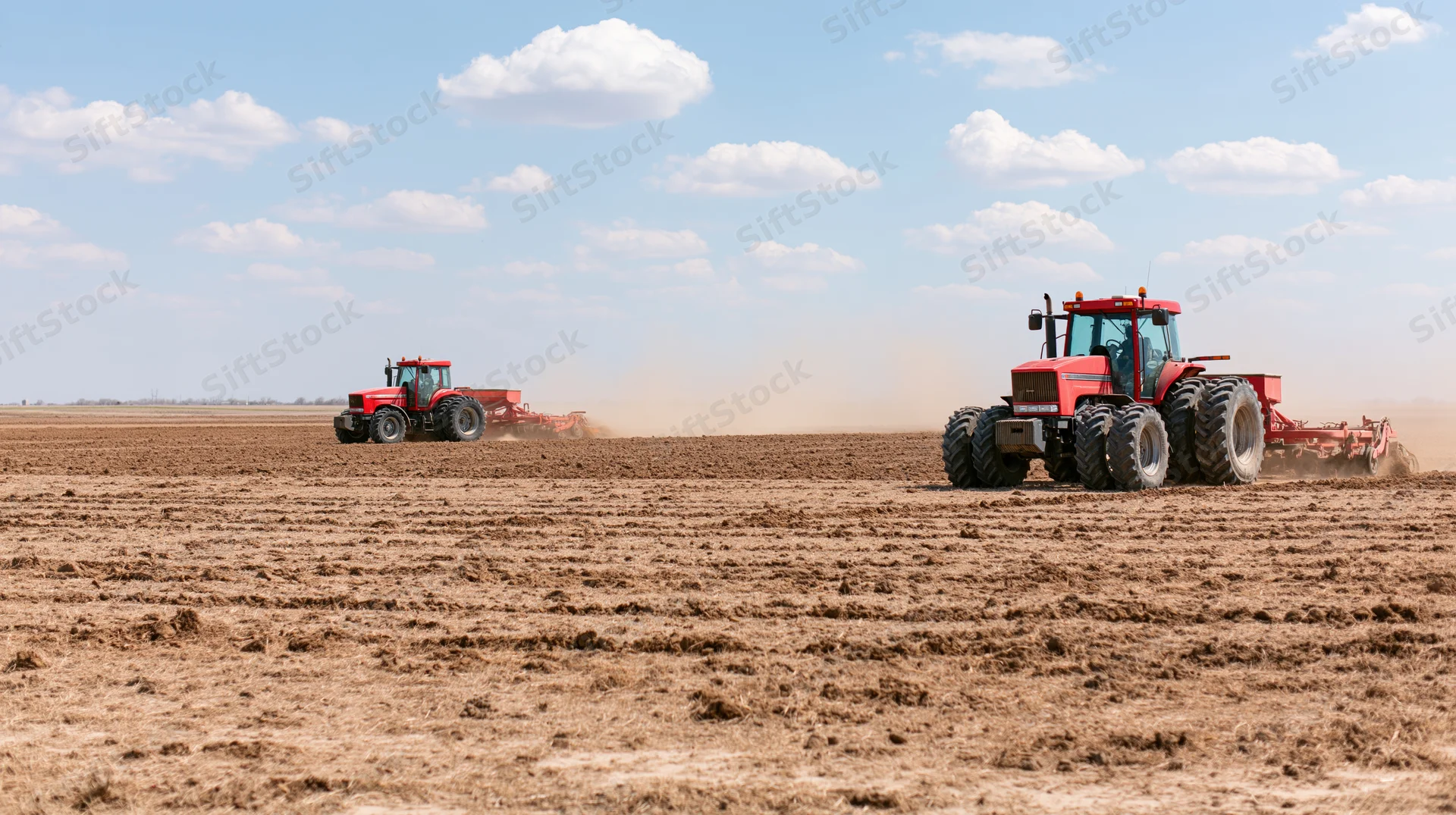 Two red tractors pulling tillage equipment across a freshly tilled field under a blue sky with scattered clouds