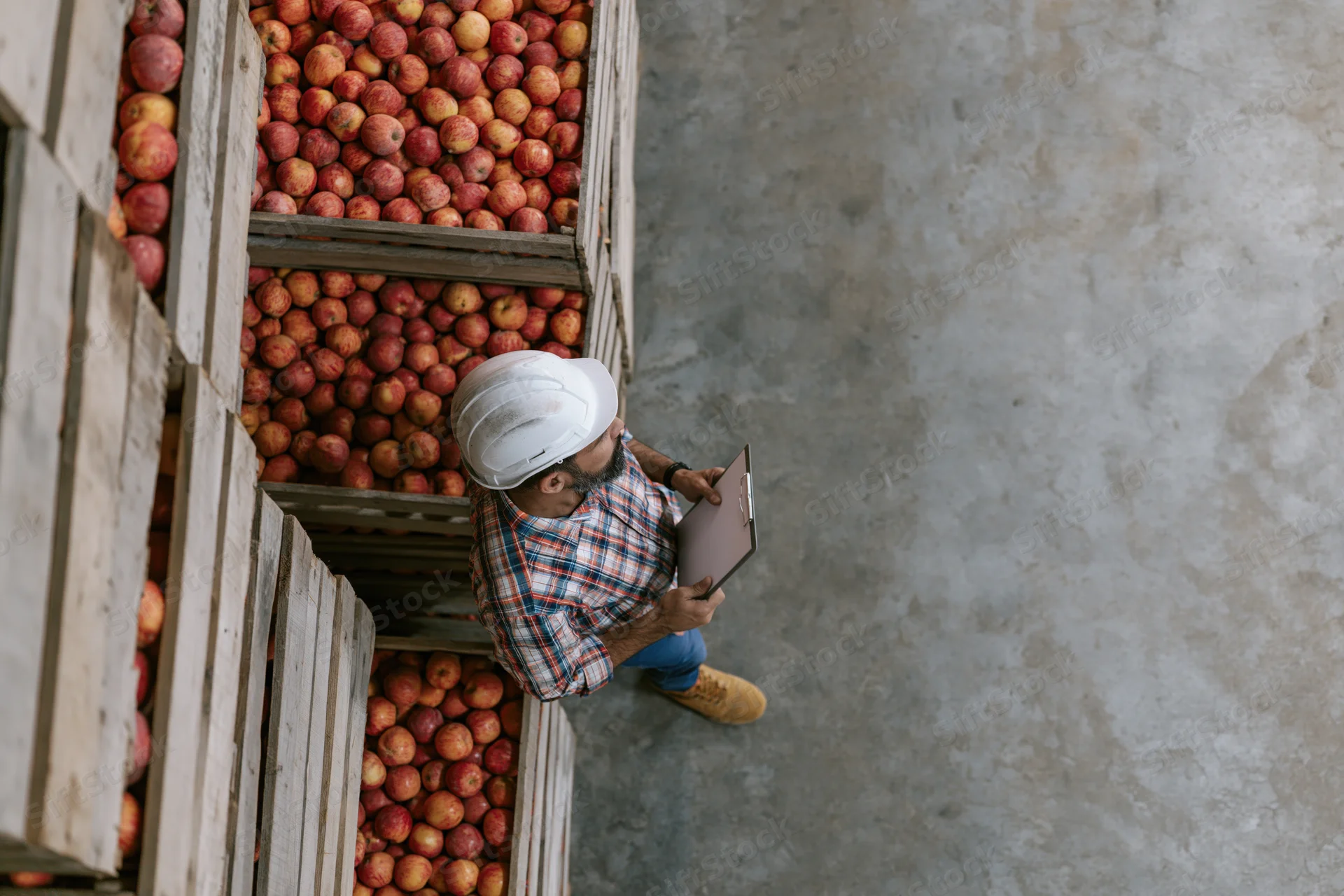 Warehouse worker in hard hat holding clipboard beside stacked crates of apples in storage facility