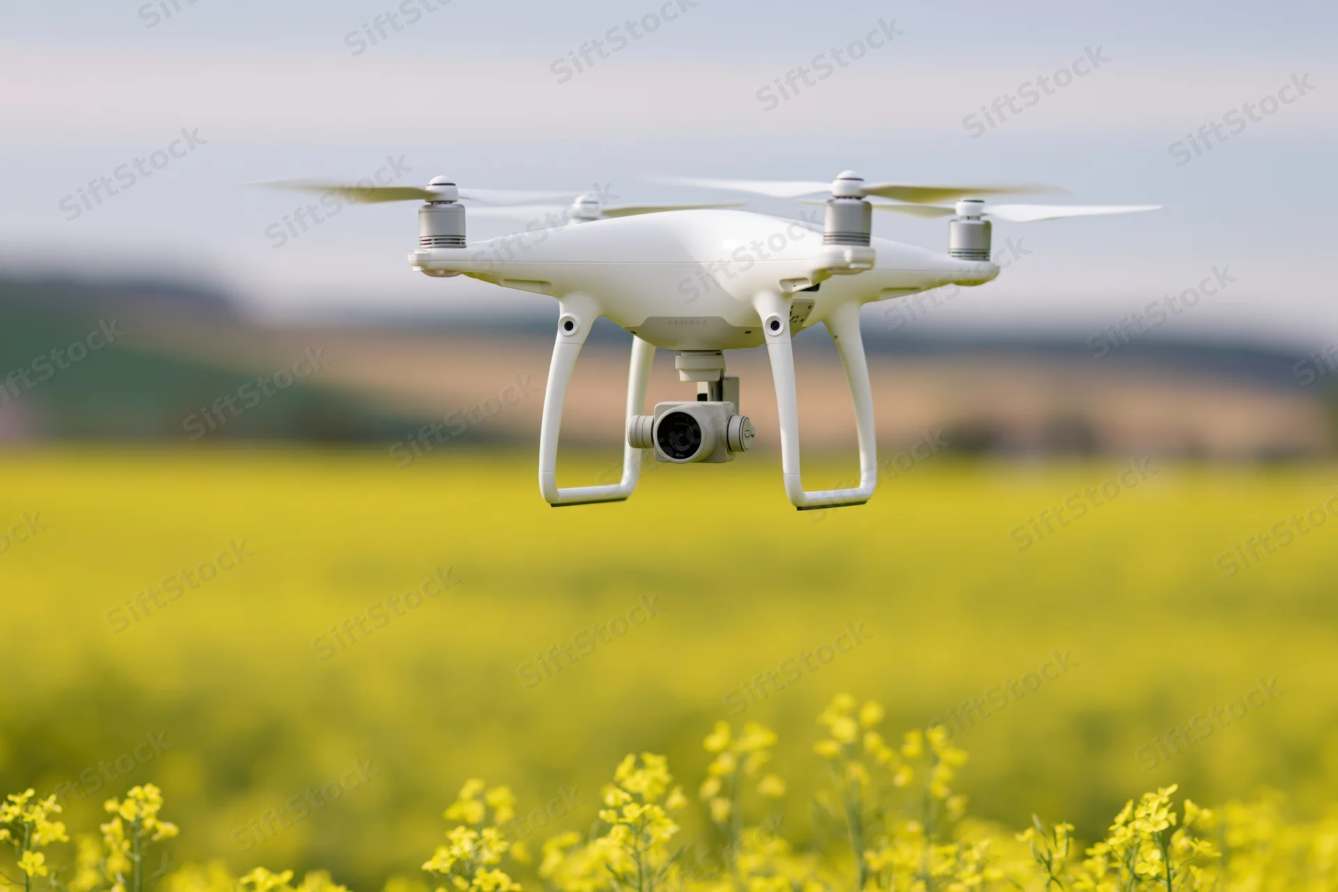 White quadcopter drone hovering over blooming canola field with camera attached