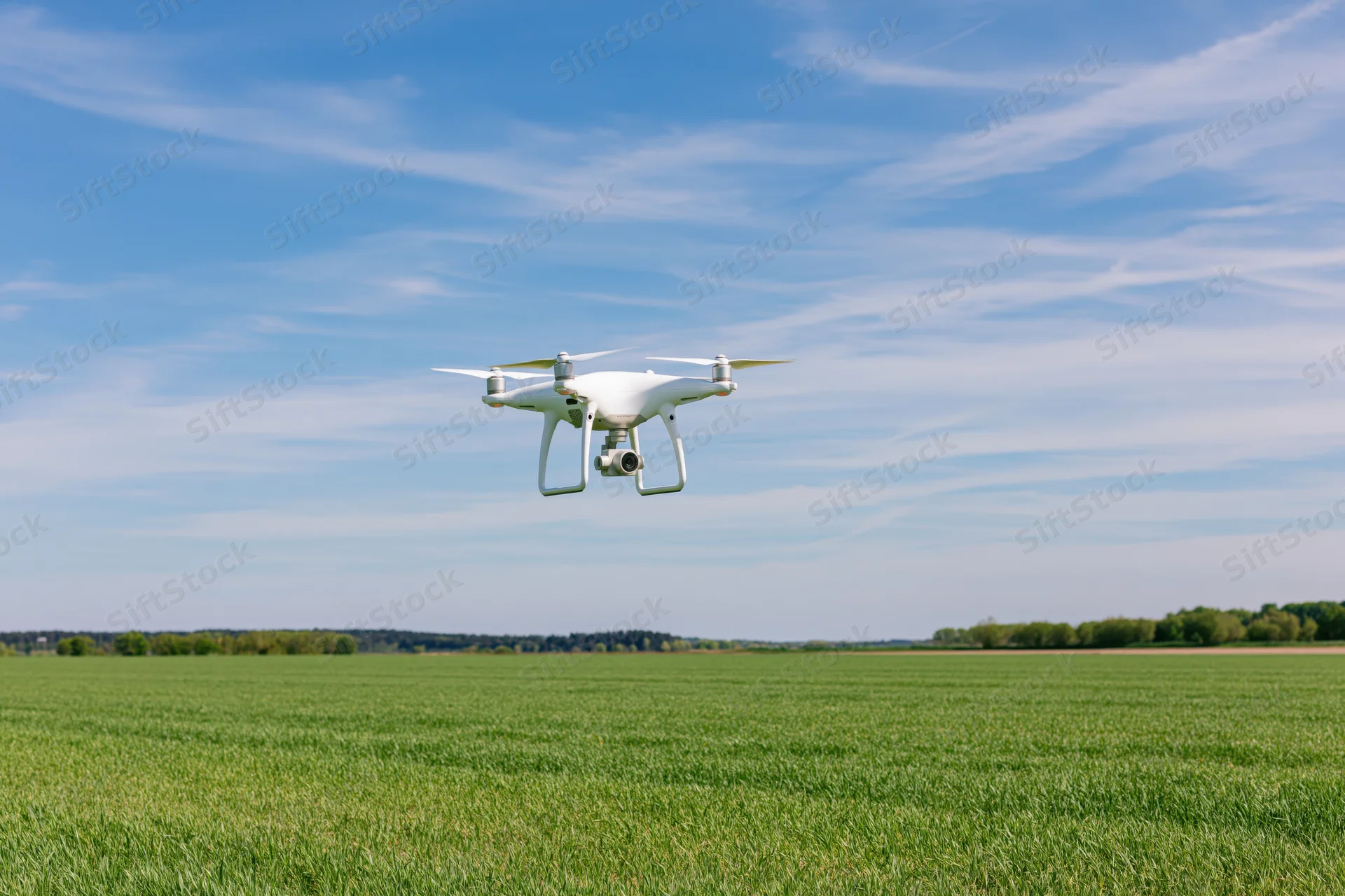 White quadcopter drone hovering over a green field under a blue sky