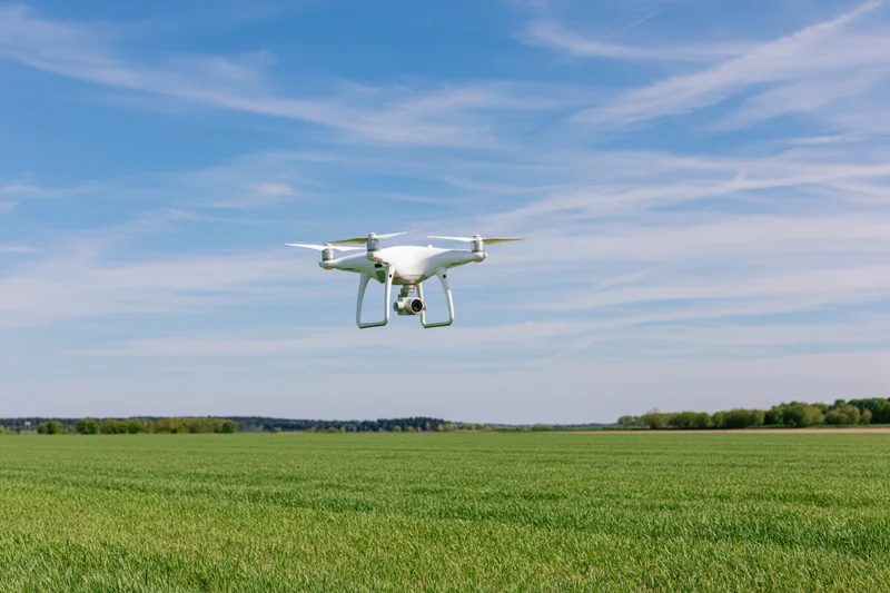 White quadcopter drone hovering over a green field under a blue sky