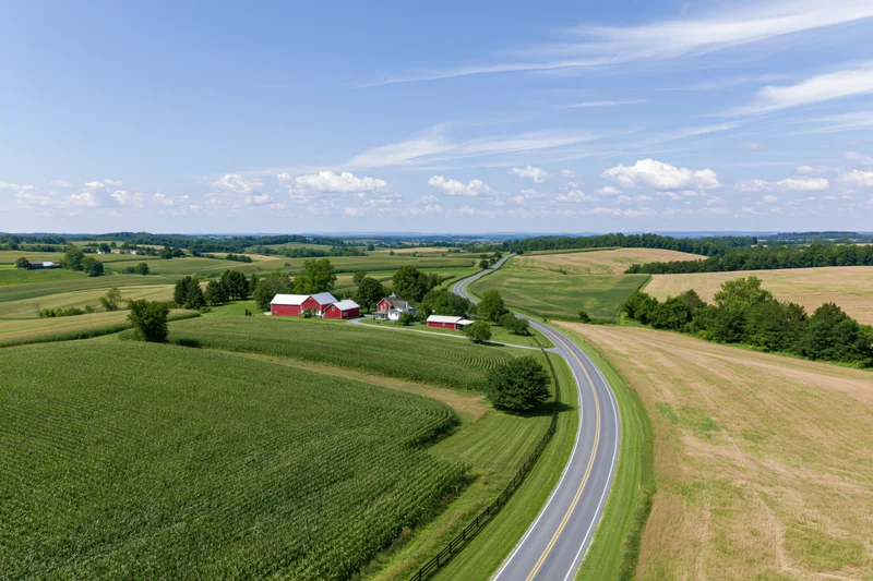 Aerial view of a rural american farm with red barns and winding road through green fields and cornfields under blue sky with clouds