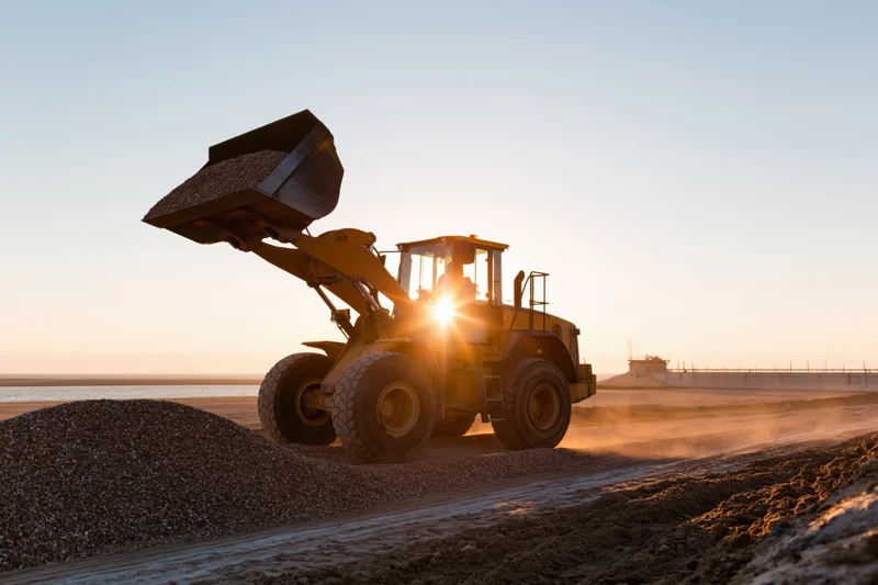Yellow wheel loader loading gravel at coastal construction site during sunset