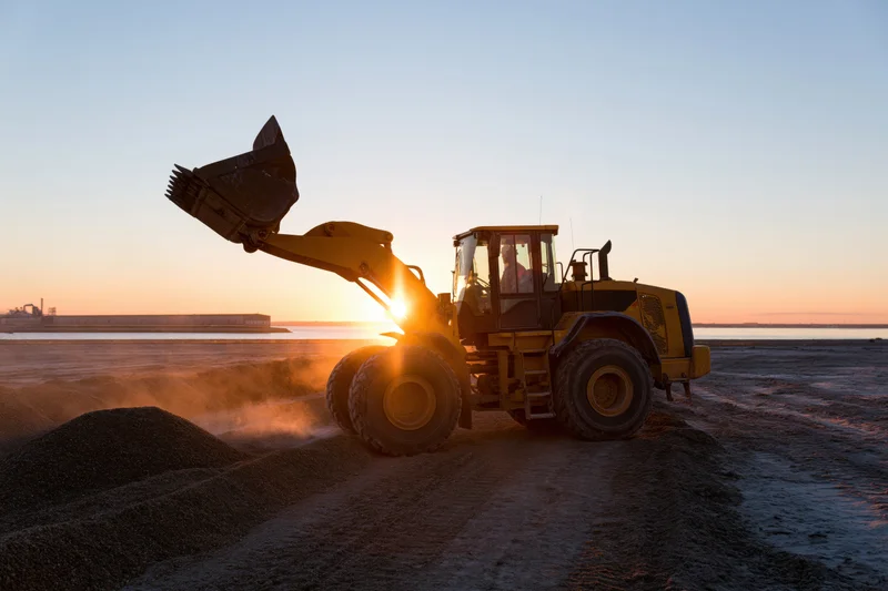 Yellow wheel loader lifting bucket at coastal construction site during sunset