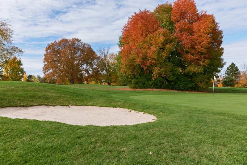 Golf course green with sand bunker and white flag pin surrounded by autumn trees
