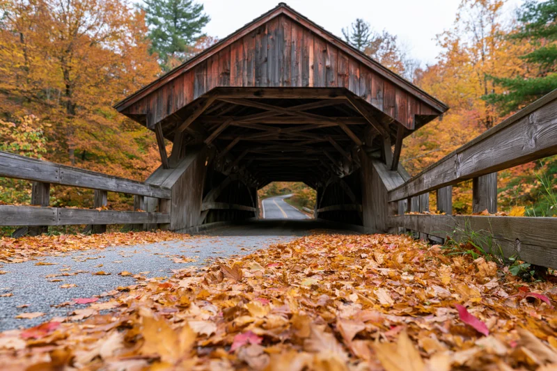 Wooden covered bridge spanning a rural road in autumn with fallen leaves on the pavement
