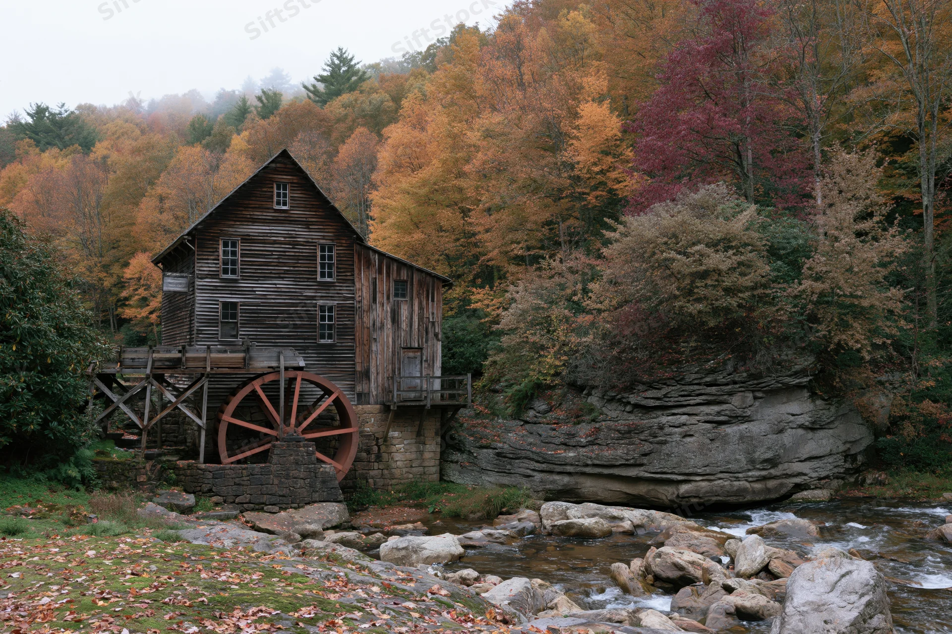 Historic wooden grist mill with rust-red waterwheel beside a rocky stream in autumn forest