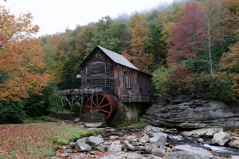 Historic wooden grist mill with rust-red waterwheel beside a rocky stream in autumn forest