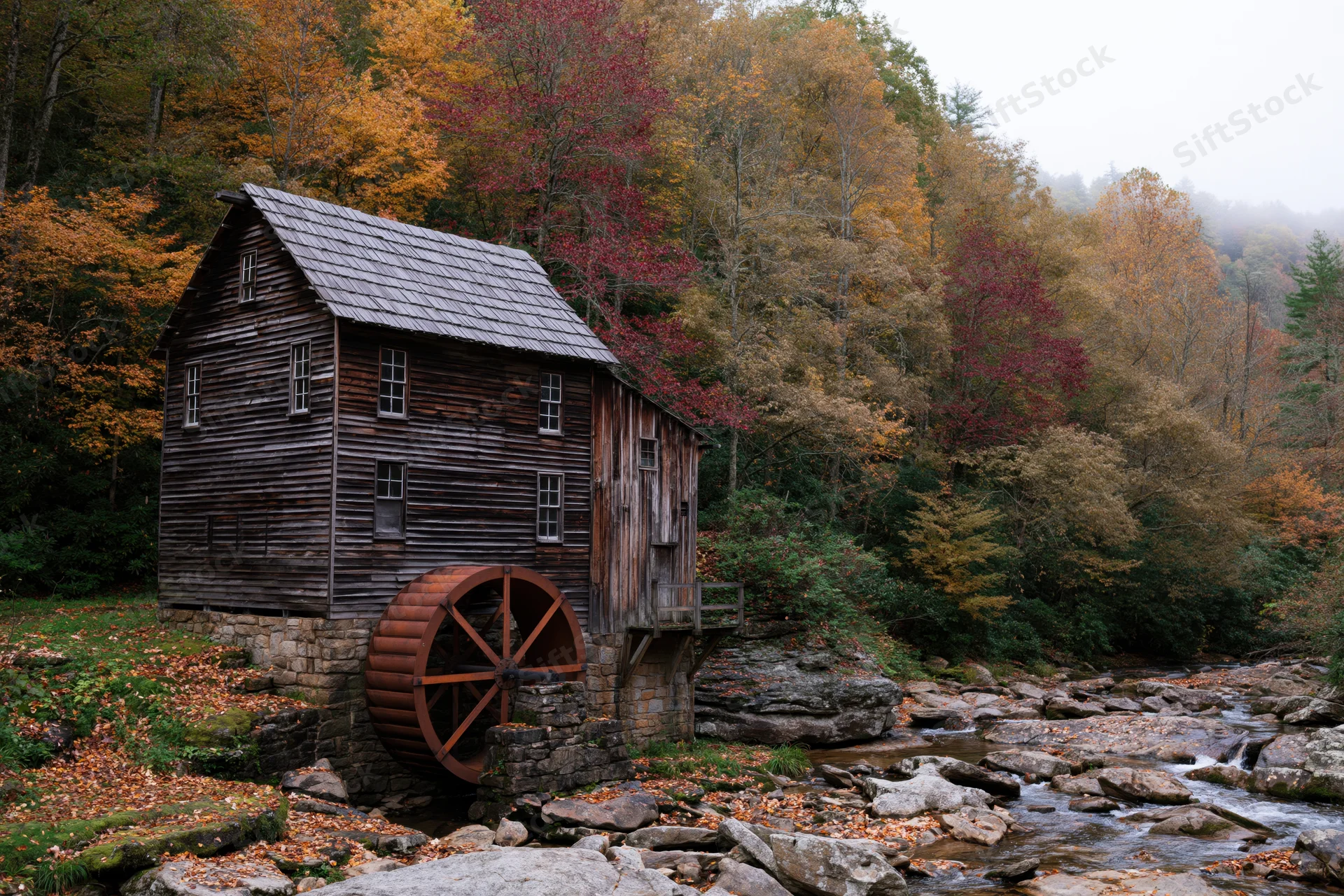 Historic wooden grist mill with rust-red waterwheel beside a rocky stream in autumn forest