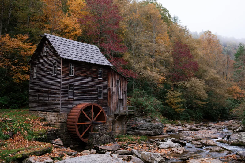 Historic wooden grist mill with rust-red waterwheel beside a rocky stream in autumn forest