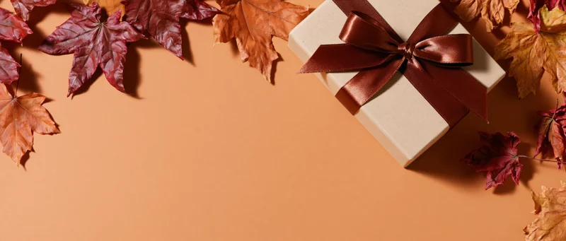 Gift box with brown satin ribbon bow surrounded by autumn maple leaves on orange background