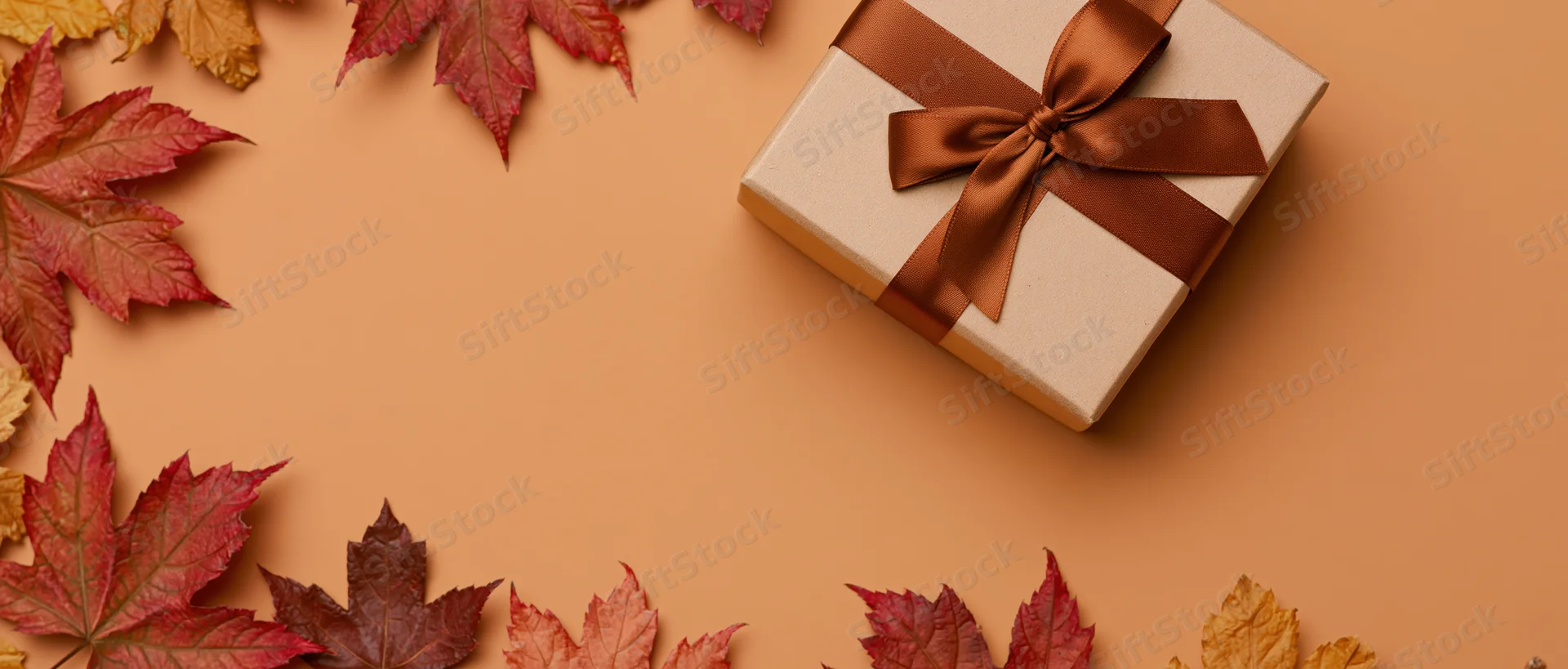 Gift box wrapped in kraft paper with brown satin ribbon bow surrounded by autumn maple leaves on warm orange background