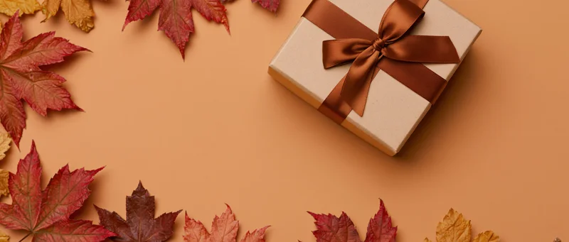 Gift box wrapped in kraft paper with brown satin ribbon bow surrounded by autumn maple leaves on warm orange background