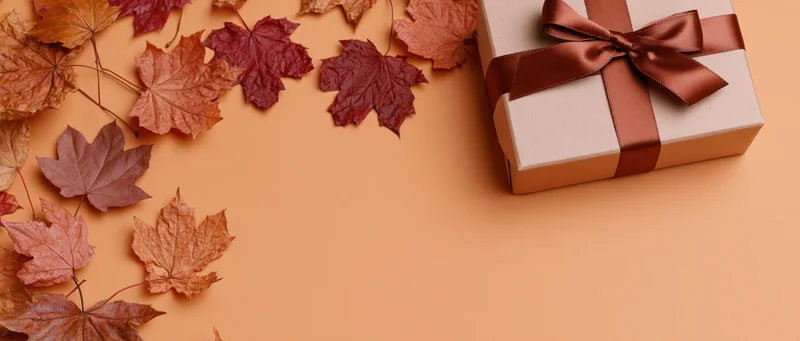 Gift box with brown satin ribbon and autumn maple leaves arranged on warm orange surface