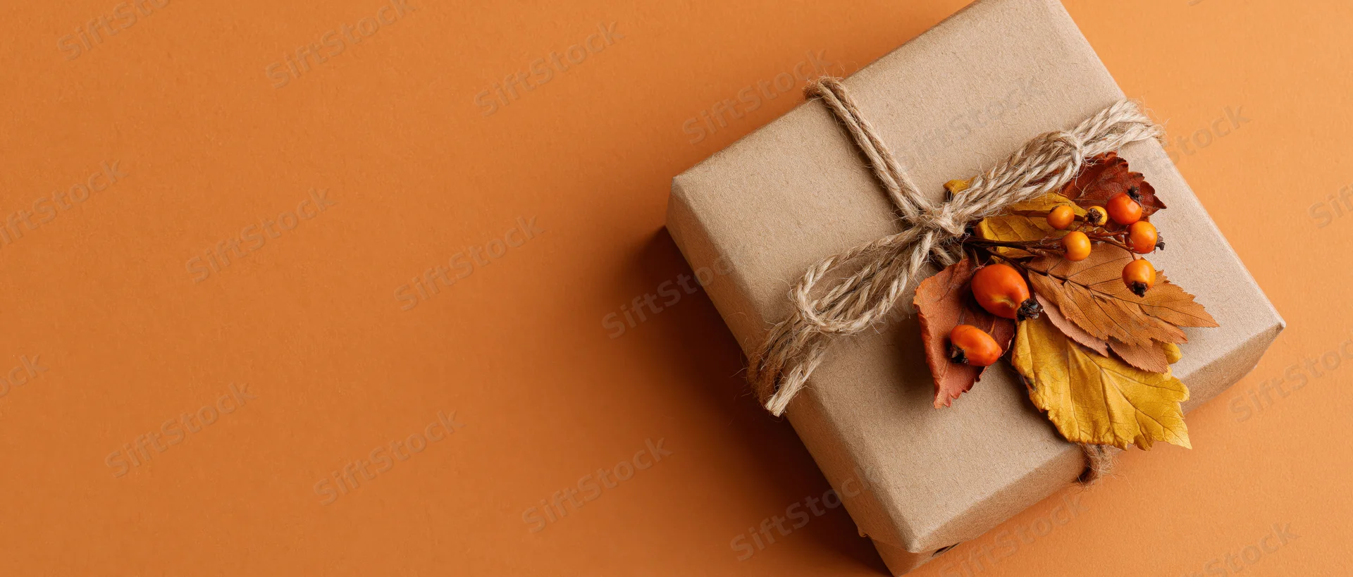 Kraft paper gift box tied with twine and decorated with dried autumn leaves and berries on orange background