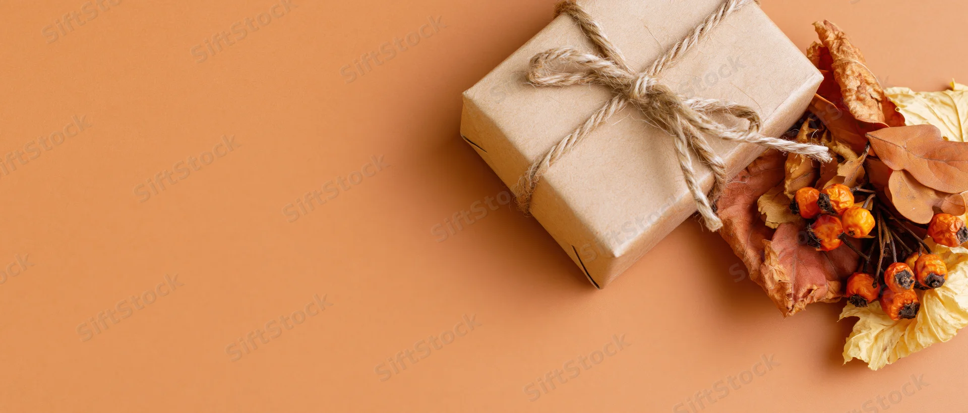 Kraft paper gift box tied with twine beside dried autumn leaves and berries on warm orange surface