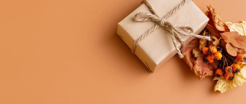 Kraft paper gift box tied with twine beside dried autumn leaves and berries on warm orange surface