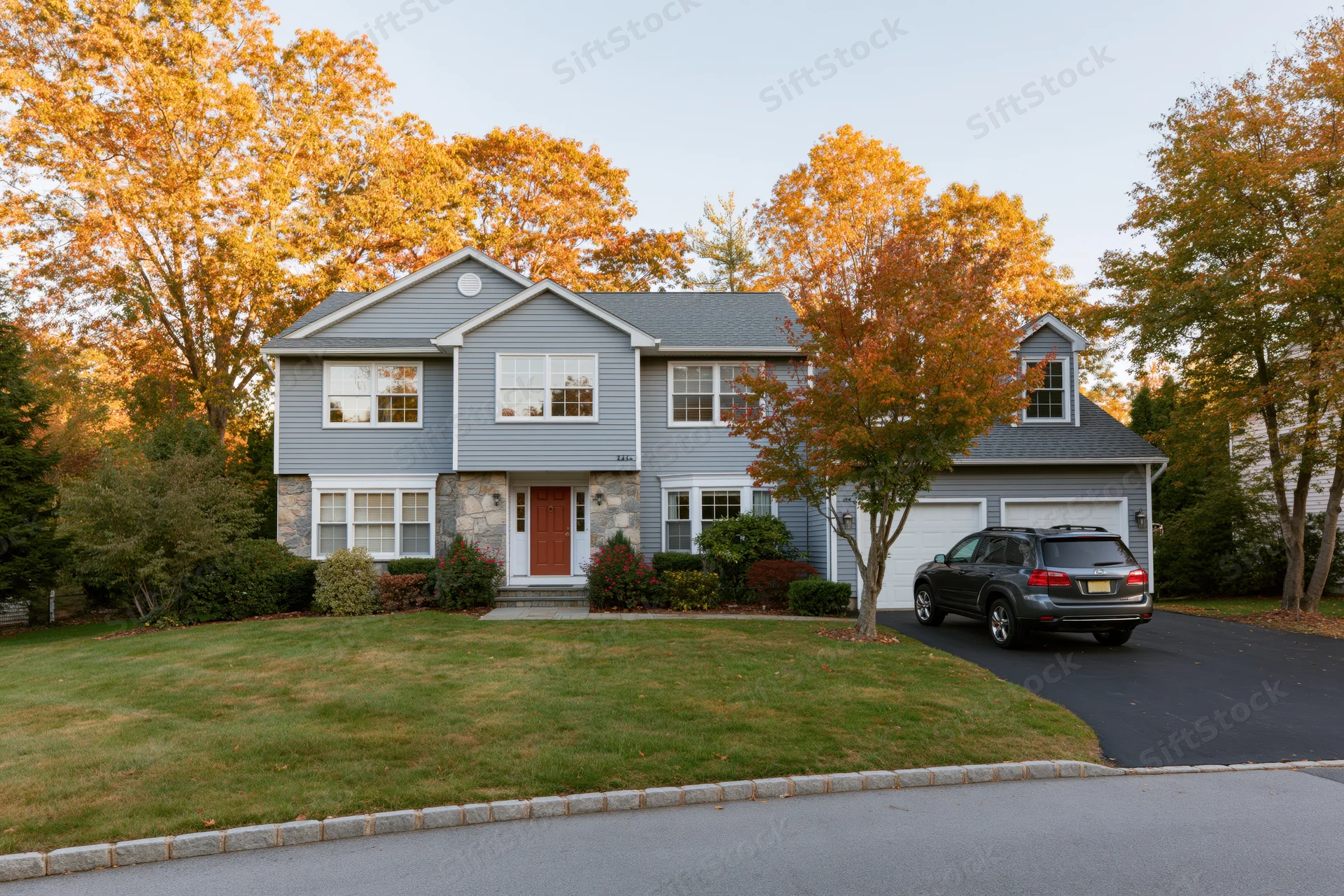Two-story suburban house with grey vinyl siding and stone accents, surrounded by autumn trees and a paved driveway with an SUV parked in front