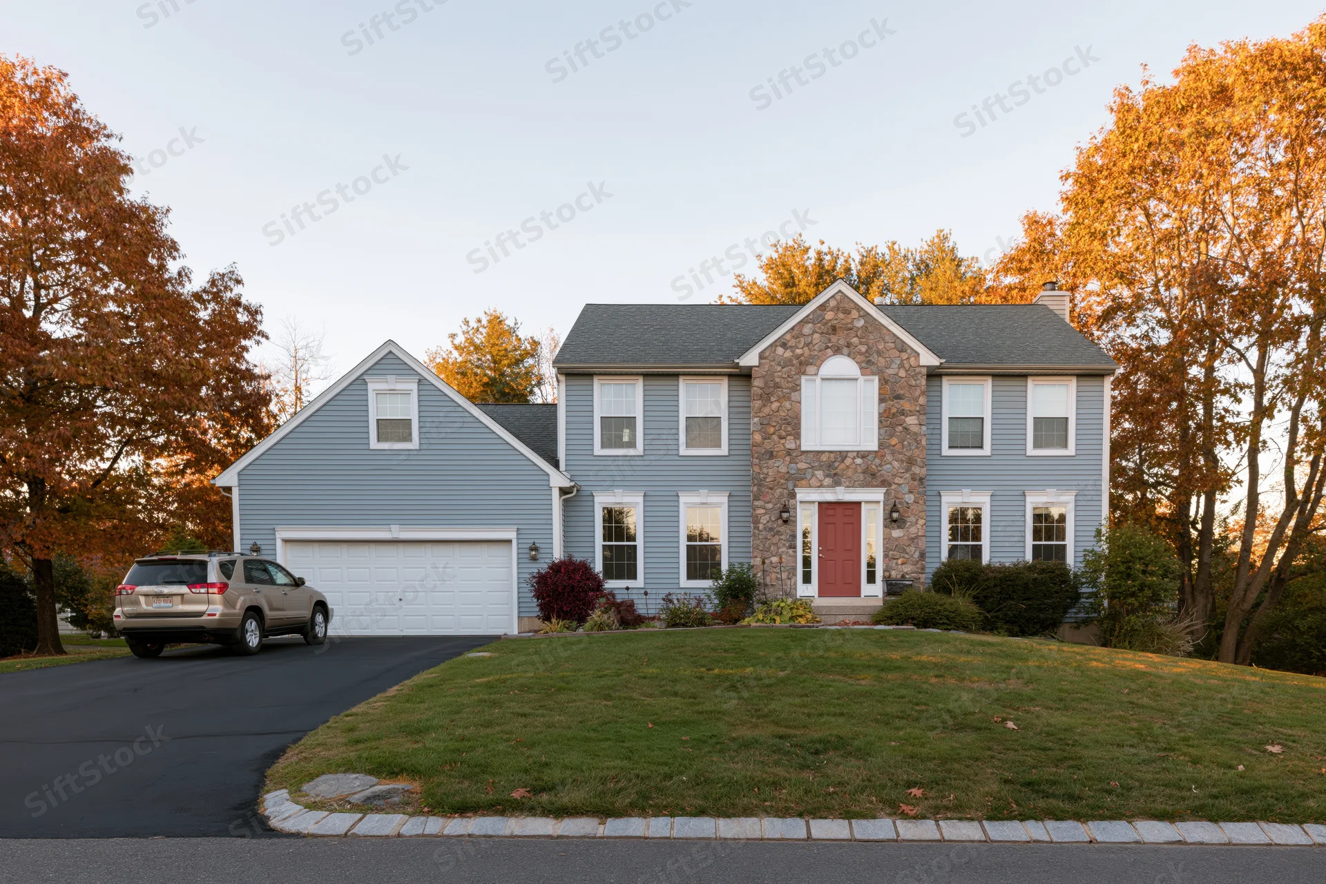 Two-story suburban house with grey vinyl siding and stone facade, red front door, driveway and garage, surrounded by autumn trees and lawn