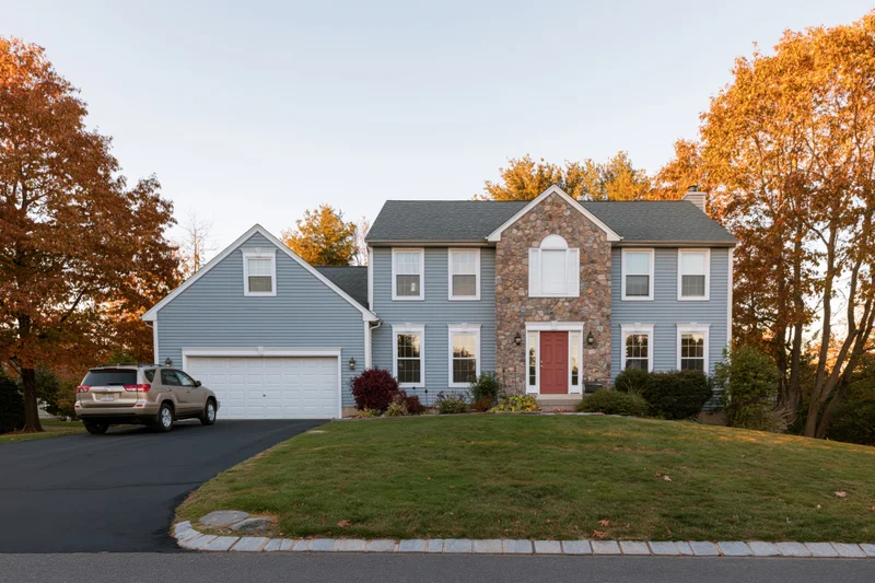 Two-story suburban house with grey vinyl siding and stone facade, red front door, driveway and garage, surrounded by autumn trees and lawn