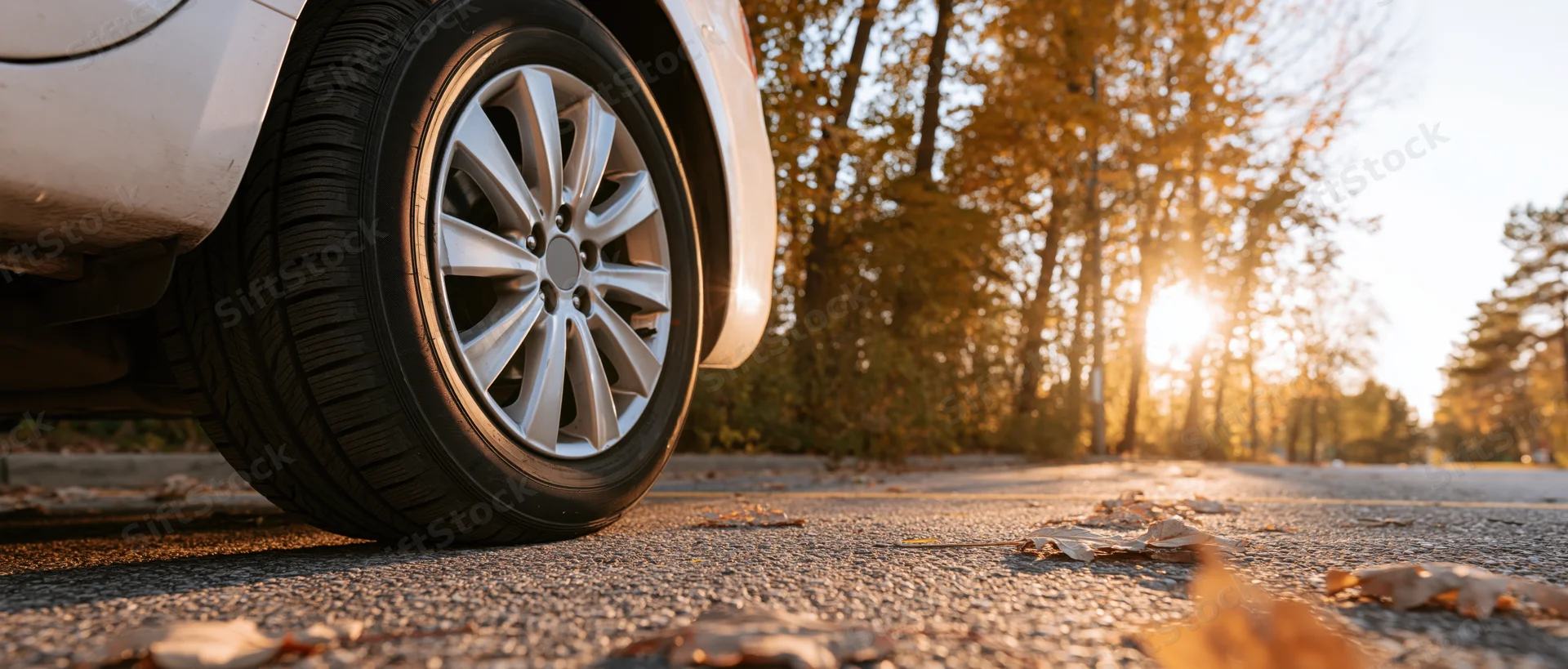 Low-angle close-up of a car's front wheel and alloy rim on a leaf-strewn road at golden hour