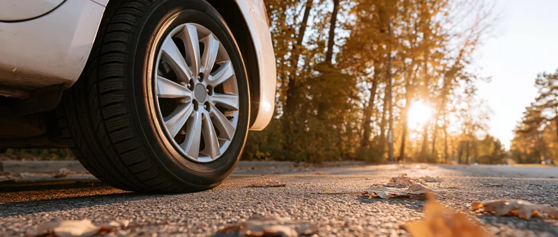 Low-angle close-up of a car's front wheel and alloy rim on a leaf-strewn road at golden hour
