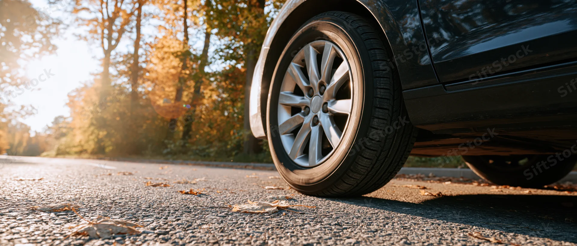 Close-up of a car's front wheel and alloy rim on a leaf-strewn road in autumn sunlight