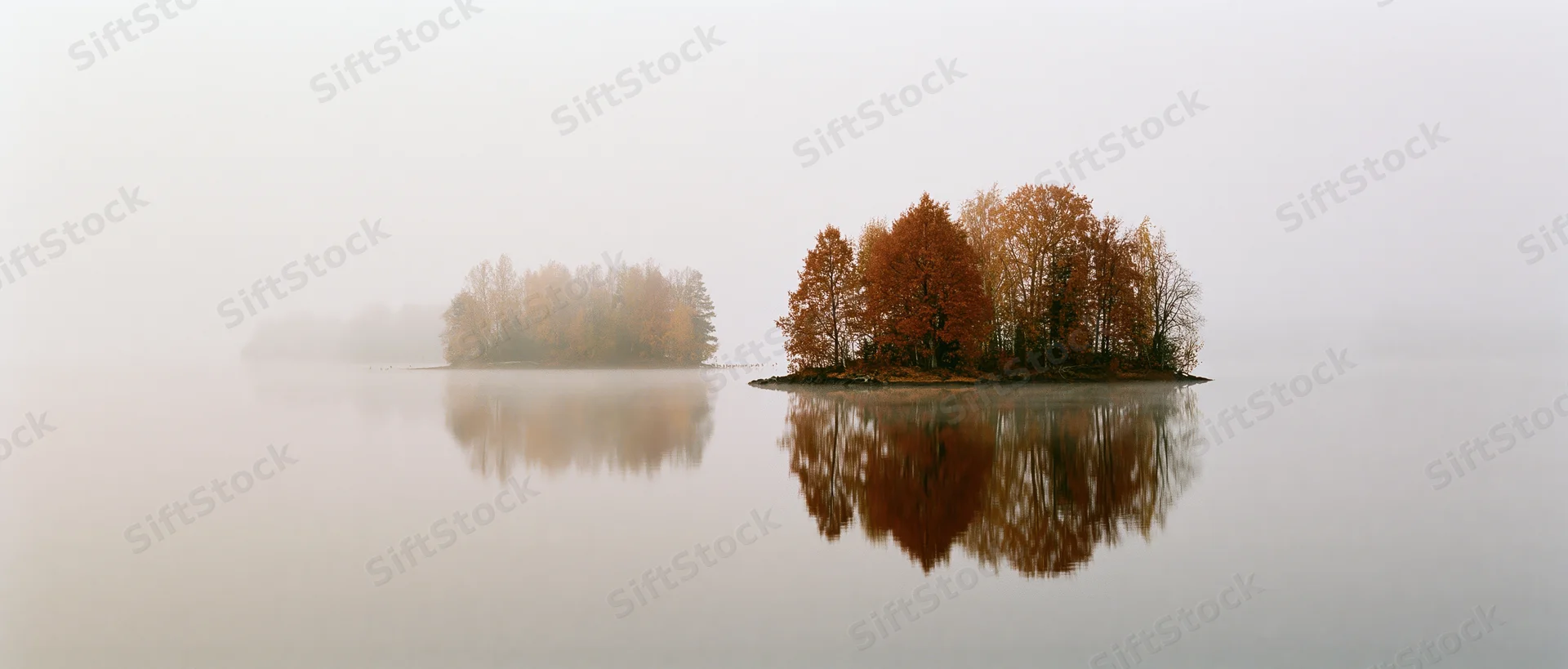 Small wooded islands with autumn trees reflected in calm misty lake at dawn